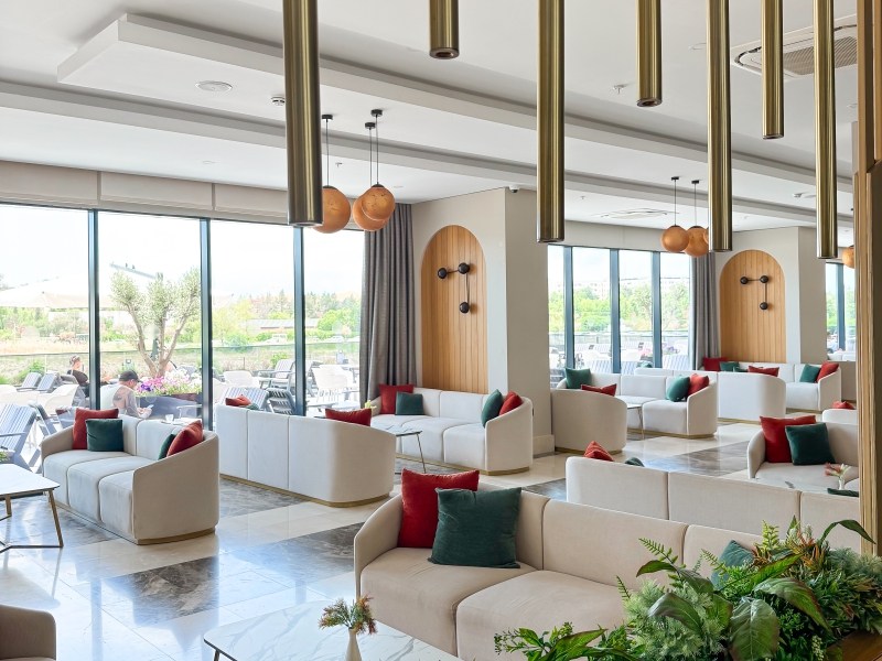 Bright, Spacious Lounge Area With White Sofas, Colorful Accent Pillows, And Large Floor To Ceiling Windows Overlooking A Terrace.
